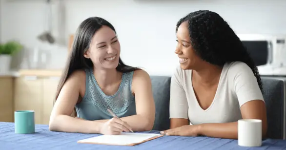 Two smiling young women sit at a table reviewing and signing paperwork together, illustrating questions to ask before signing a composite company contract.