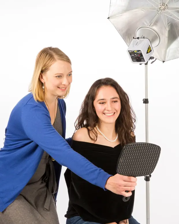 A photographer in a blue sweater holds up a handheld mirror for a smiling young woman seated for her portrait, with a studio light and silver umbrella beside them.