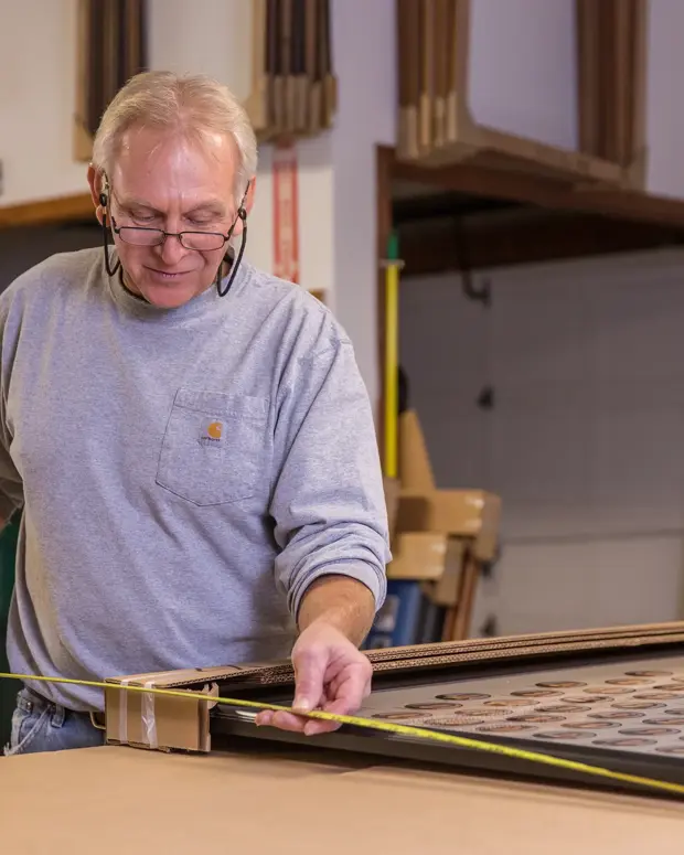 A man in a gray Carhartt shirt uses a tape measure to check the dimensions of a framed composite of oval portraits in the workshop.