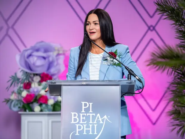 Woman standing on a stage at a podium with Pi Beta Phi on it giving a speech. 