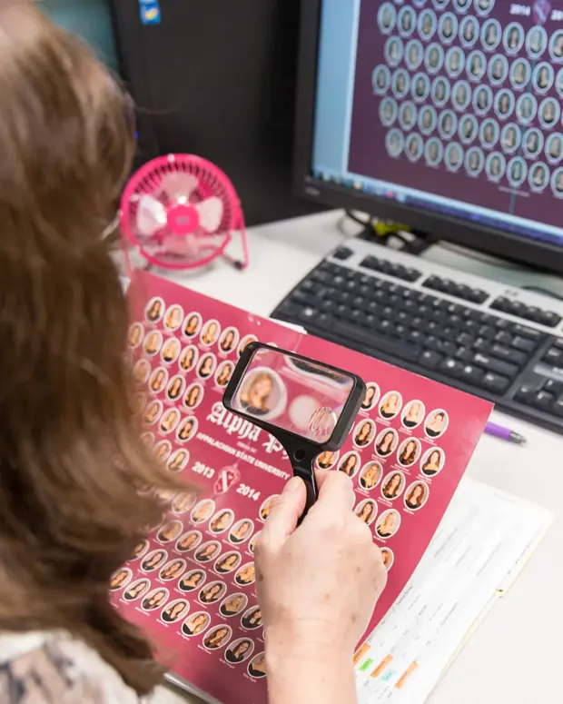 Over-the-shoulder view of a woman using a magnifier to inspect a maroon composite proof of sorority portraits, with the same layout shown on her monitor.
