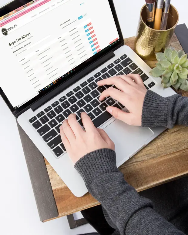 Overhead view of a person's hands typing on a laptop that displays the Digital Pix Composites planner with a sign-up sheet of member names, emails, and scheduled times.