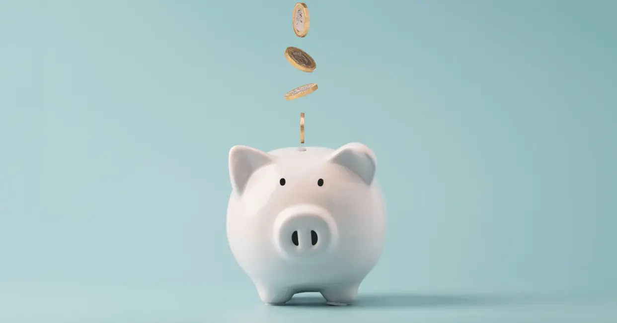 A white ceramic piggy bank with gold coins dropping into it on a blue background, symbolizing the cost of a Greek life composite.