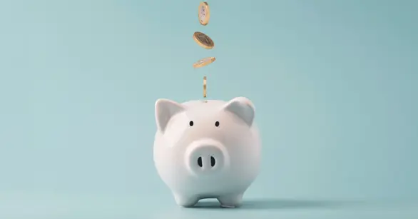 A white ceramic piggy bank with gold coins dropping into it on a blue background, symbolizing the cost of a Greek life composite.
